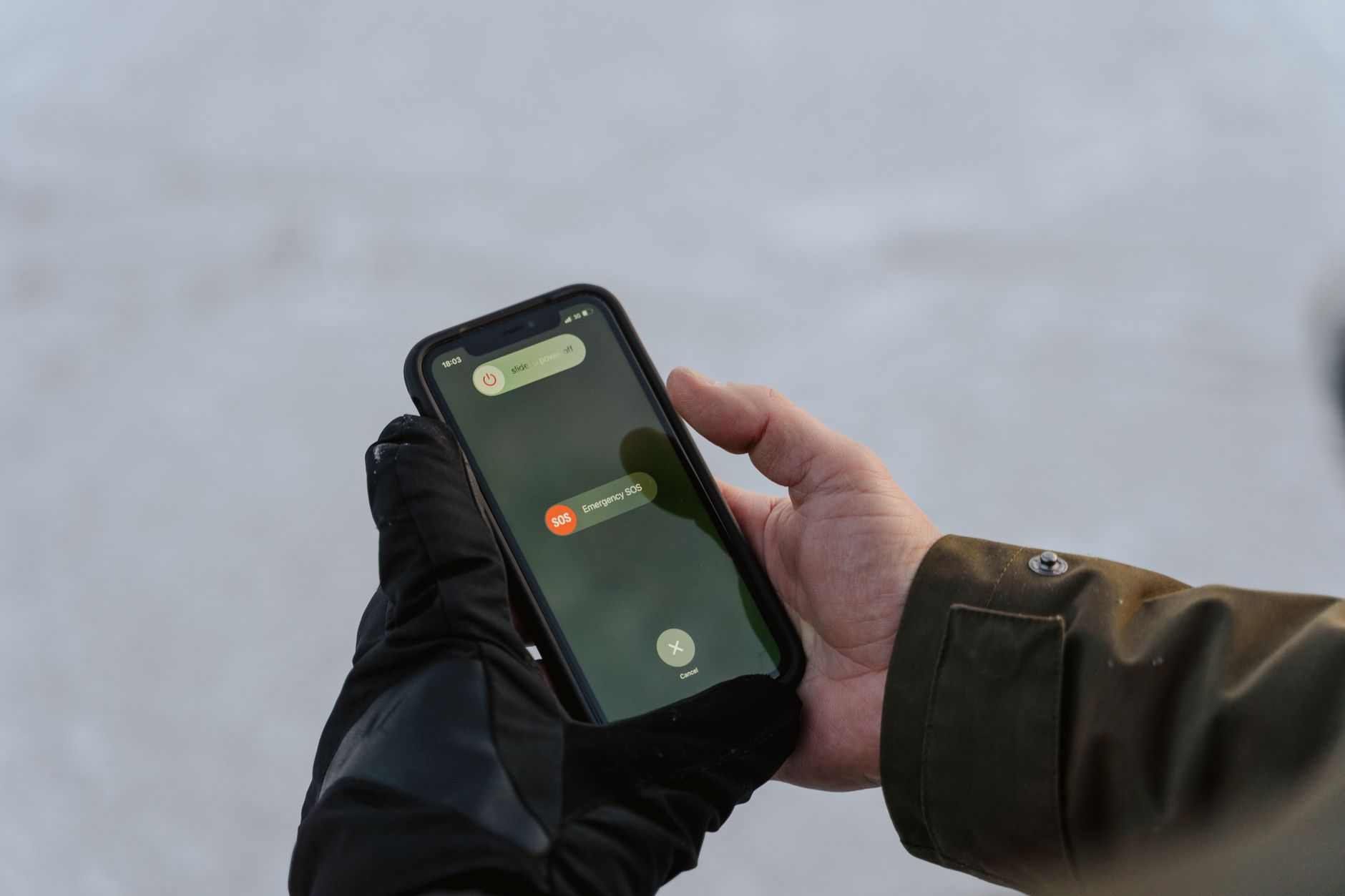 Osoba w czarnych rękawiczkach trzymająca telefon z otwartą aplikacją SOS na ekranie, na tle zasnężony krajobraz.