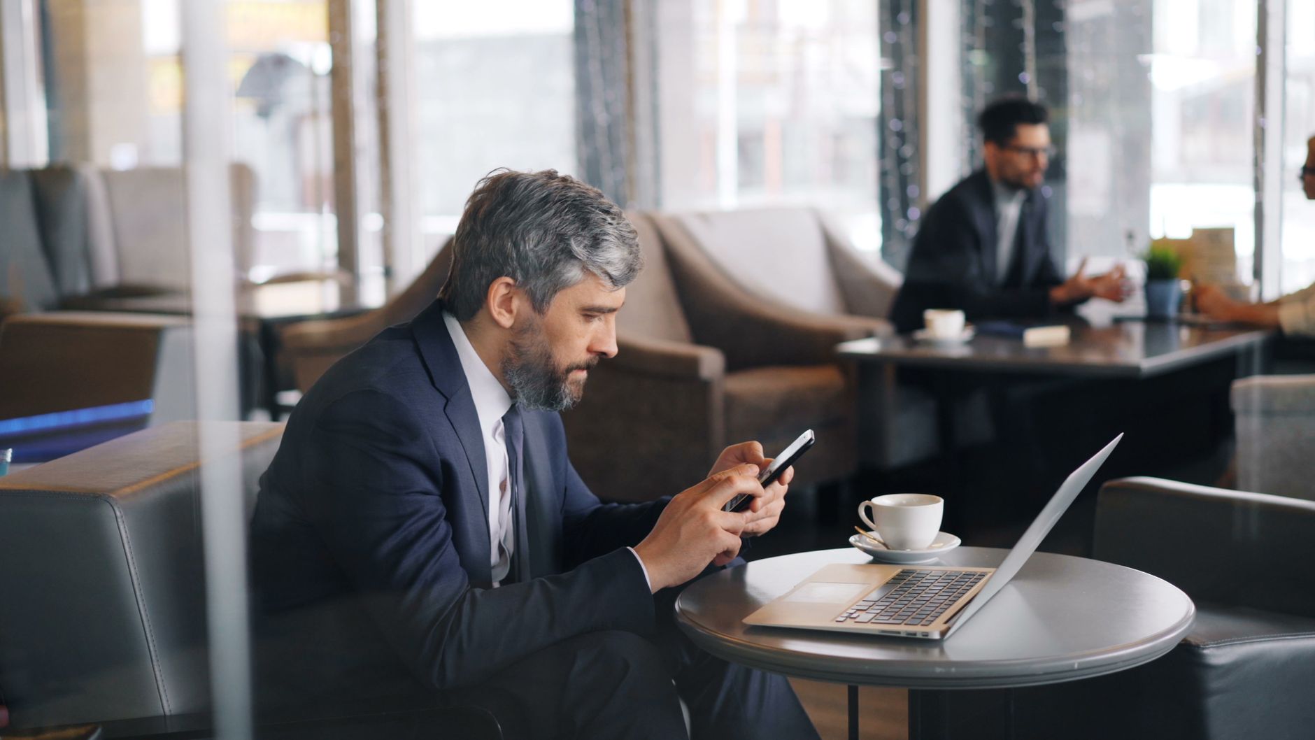 Businessman in dark suit sitting at round table with laptop and coffee, holding smartphone in modern office