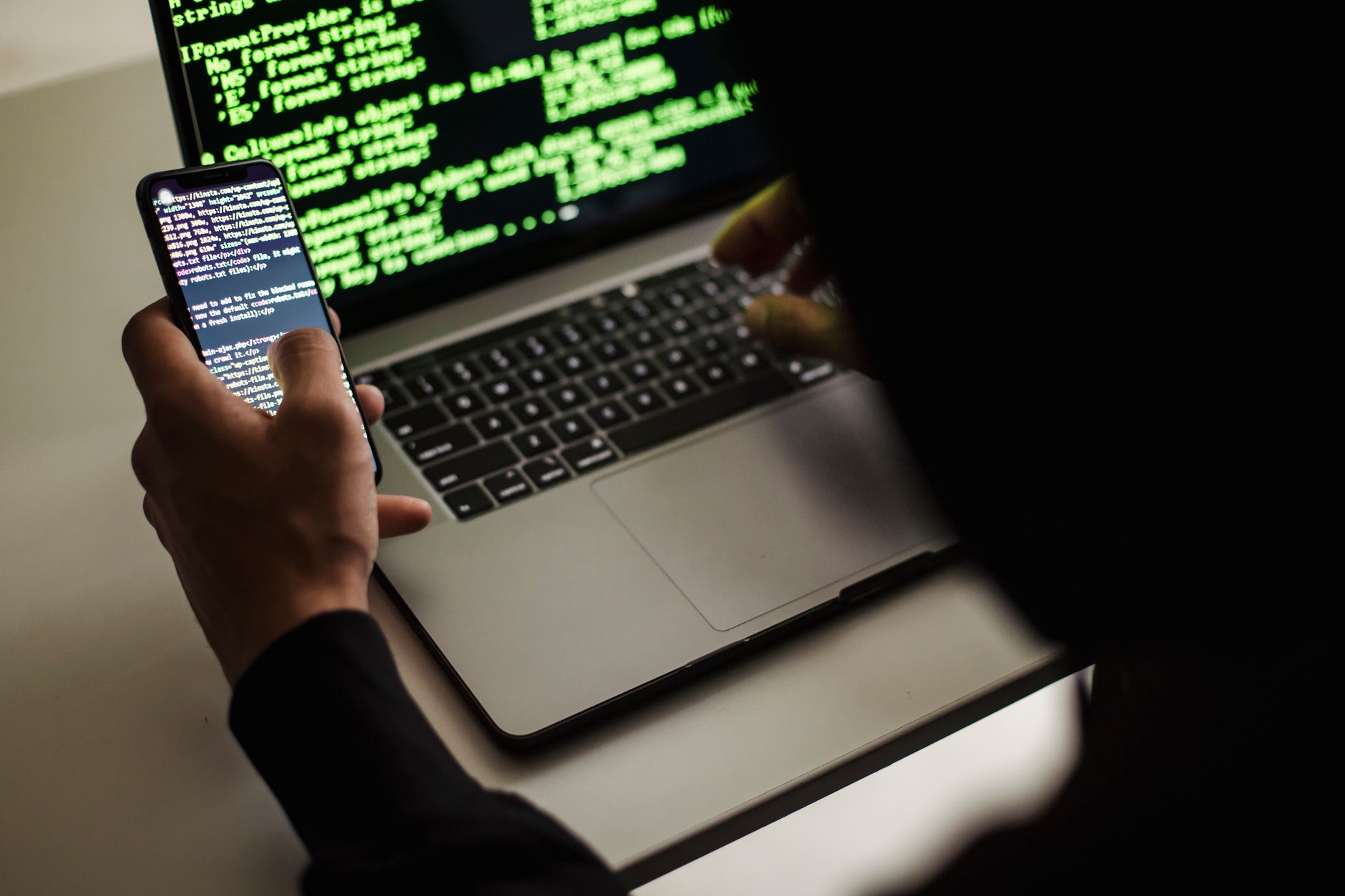 Person holding smartphone displaying code while working at laptop with green terminal text visible on screen