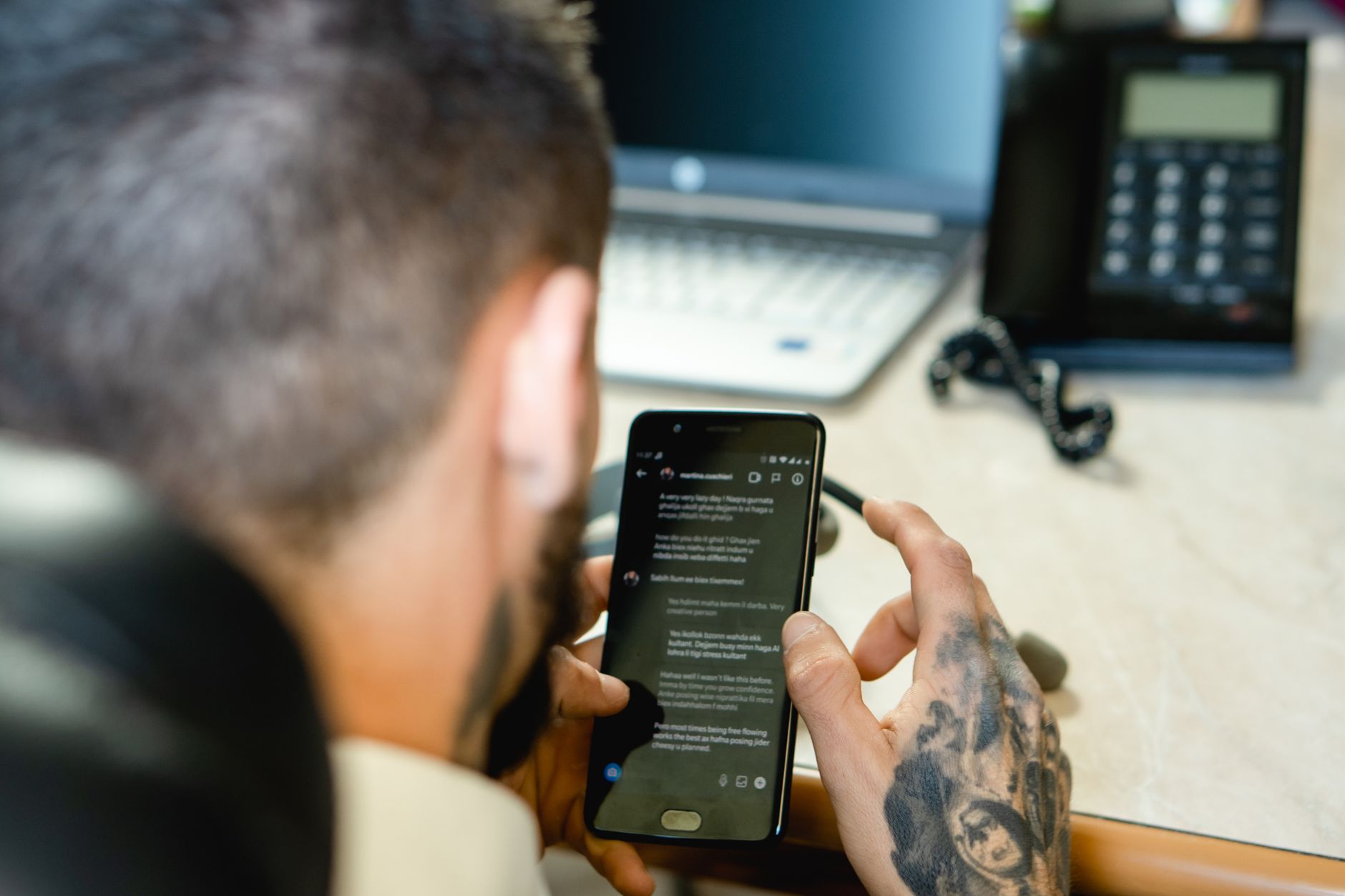 Person with tattooed hand holding smartphone displaying text content at office desk with laptop and phone visible in background
