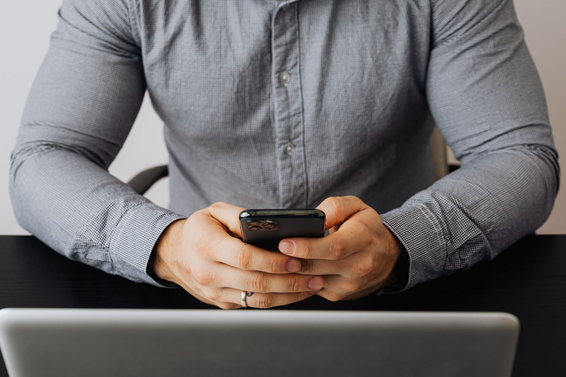 Man in gray button-up shirt holding smartphone while seated at desk with laptop
