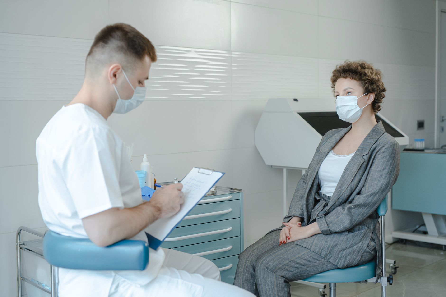 Młody sanitariusz w białym ubraniu i masce rozmawia z pacjentką w szarej marynarce, trzymając schowek w nowoczesnym gabinecie medycznym.
